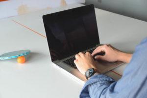 Ihminen kirjoittaa kannettavalla tietokoneella pingispöydän päällä. Pöydällä pingismaila ja -pallo.