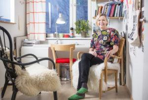 Tekstiilitaiteilija Laura Isoniemi istuu puisella tuolilla värikkään huoneen nurkassa.