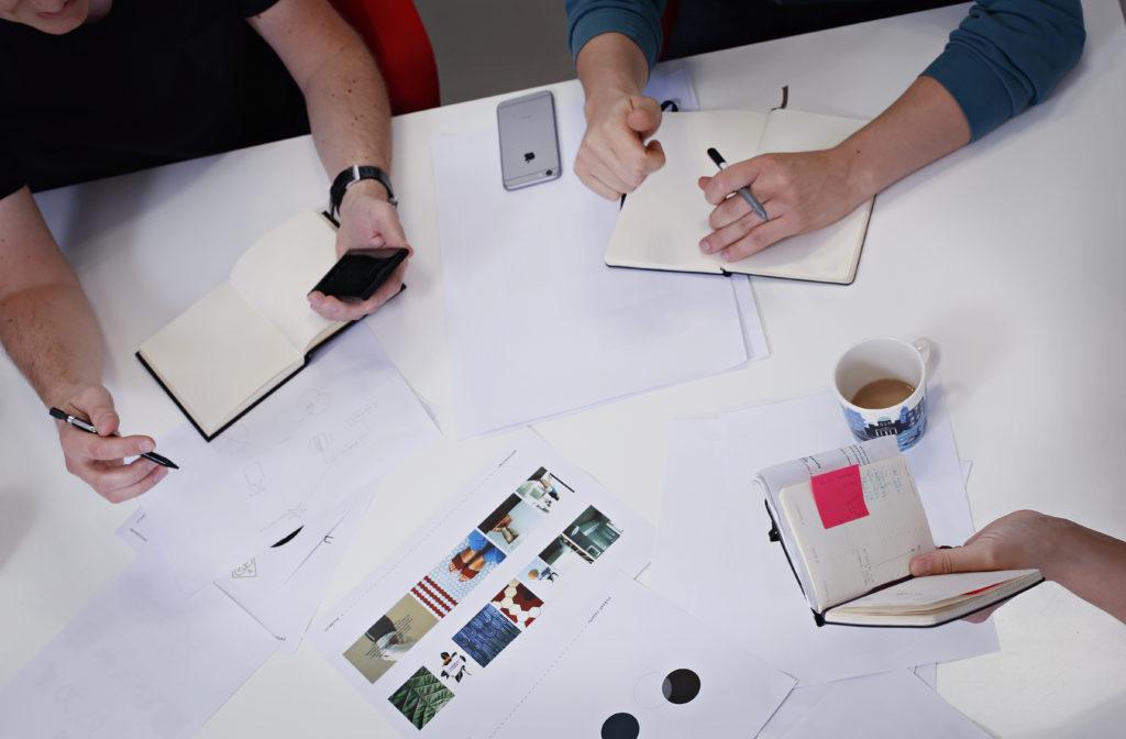 Suunnittelijat työpöydän ääressä, pöydällä vihkoja ja suunnitelmapapereita